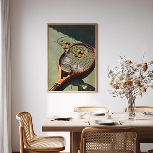 May include: A framed print depicting a tennis racket with two martini glasses containing olives. The artwork has a vintage style, with green and brown tones. The image is displayed above a wooden dining table.