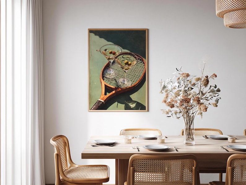 May include: A framed print depicting a tennis racket with two martini glasses containing olives. The artwork has a vintage style, with green and brown tones. The image is displayed above a wooden dining table.