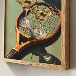 May include: Framed print of two martini glasses with olives on a vintage tennis racket. The artwork has a light green background and a wooden frame, creating a classic and stylish aesthetic.