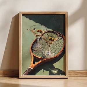 May include: A framed art print depicting two martini glasses with olives, placed on a vintage tennis racket. The artwork has a green and brown colour scheme, with a wooden frame. The glasses are filled with a clear liquid and garnished with olives.