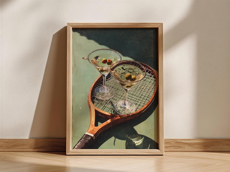 May include: A framed art print depicting two martini glasses with olives, placed on a vintage tennis racket. The artwork has a green and brown colour scheme, with a wooden frame. The glasses are filled with a clear liquid and garnished with olives.
