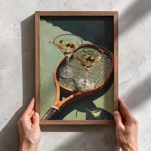May include: A framed print depicting two martini glasses with olives, placed on a vintage tennis racket. The artwork has a green and brown colour scheme, with a retro aesthetic. The print is held by two hands.