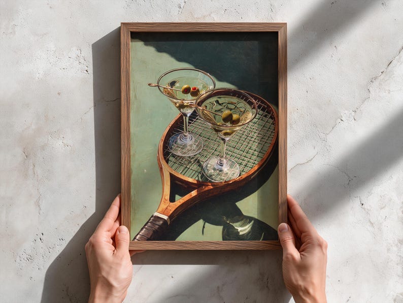 May include: A framed print depicting two martini glasses with olives, placed on a vintage tennis racket. The artwork has a green and brown colour scheme, with a retro aesthetic. The print is held by two hands.