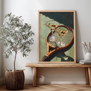 May include: A framed print depicting two martini glasses with olives, placed on a vintage tennis racket. The artwork is displayed on a wooden bench, with a potted plant and a vase. The color scheme includes greens, browns, and whites.