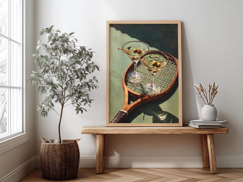 May include: A framed print depicting two martini glasses with olives, placed on a vintage tennis racket. The artwork is displayed on a wooden bench, with a potted plant and a vase. The color scheme includes greens, browns, and whites.