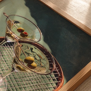 May include: Two martini glasses with olives on a decorative tray. The glasses are filled with a clear liquid and garnished with green olives. The tray has a copper-coloured rim and a grid pattern.