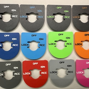 May include: A collection of twelve U-shaped ignition switch plates in various colors including gray, blue, green, orange, red, and pink. Each plate features a central cutout and the printed text labels OFF, IGN, LOCK, and ACC in contrasting colors.