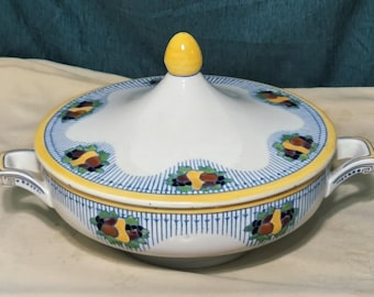 May include: A white ceramic serving dish with a lid, featuring a yellow finial. The dish has a blue and yellow rim with floral patterns. It includes two handles and is designed for serving food.