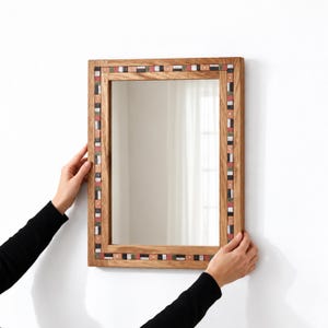 Op de afbeelding: Een rechthoekige wandspiegel met een lichtbruin houten frame. Het frame is versierd met een ingelegde rand van kleine, afwisselende rechthoekige tegeltjes in tinten van gedempt groen, wit, zwart en koper, gerangschikt in een herhalend patroon.