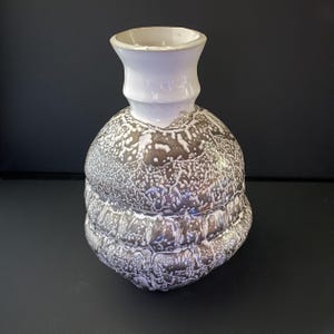 May include: A ceramic vase with a bulbous, multi-tiered body featuring a textured, metallic brown and white drip glaze. The neck is smooth, cylindrical, and finished in a solid white glaze, contrasting with the complex, cratered surface below.