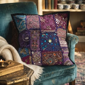 May include: A square decorative pillow featuring a patchwork design in shades of purple, blue, and teal. The surface is embellished with intricate embroidery, small mirrors, beads, and sequins arranged in various geometric and floral patterns.