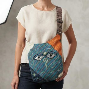 May include: A crossbody bag featuring a patchwork design with teal, orange, and cream fabric panels. The bag displays an embroidered face motif with stylized eyes and a nose, accented by thin, swirling thread lines and a striped adjustable shoulder strap.