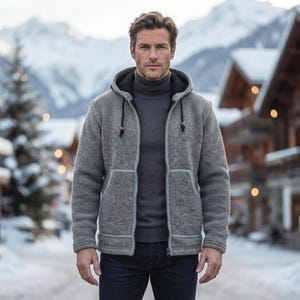 May include: A grey knit zip-up hoodie with a black interior lining and black drawstring cords, worn over a dark grey turtleneck sweater and dark blue denim jeans. The hoodie features a front kangaroo pocket and light grey trim along the zipper.