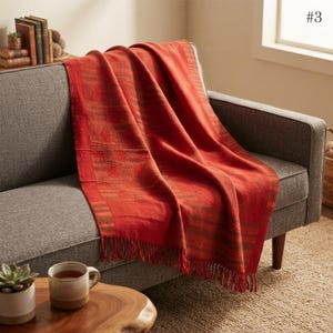 May include: A woven throw blanket in a vibrant burnt orange hue featuring subtle horizontal stripes and a fringed edge. The blanket is draped over a gray upholstered sofa, showcasing a textured fabric construction with visible vertical weave patterns.