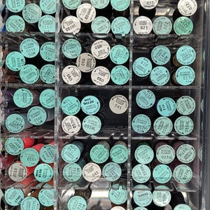 May include: A clear plastic storage organizer filled with numerous spools of Kreinik metallic blending filament. Each spool features a white circular label displaying the brand name, color code, and the text 50M, arranged in a grid pattern.