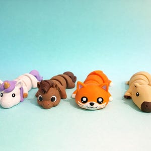 Könnte beinhalten: Vier handgefertigte Tierfiguren aus Ton: ein Einhorn, ein Pferd, ein Fuchs und ein Capybara. Das Einhorn hat eine lila Mähne und ein Horn. Das Pferd ist braun. Der Fuchs ist orange mit weißem Gesicht. Das Capybara ist hellbraun. Alle liegen.