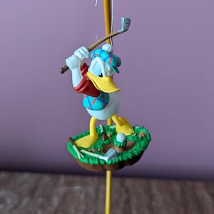 May include: A Donald Duck figurine in a golfing pose. The figure is wearing a golf outfit and a plaid hat. The figurine is on a small green base with a golf ball and club. The background is a solid purple color.