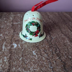 Może przedstawiać: Kremowa ozdoba choinkowa w kształcie dzwonka z czerwoną wstążką. Ozdoba posiada zielony wieniec z czerwonymi kokardkami oraz napis "Home For The Holidays". Świąteczna dekoracja.