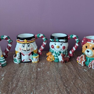 May include: Four ceramic Christmas mugs with character designs. The mugs feature Santa Claus, a Nutcracker, a snowman, and a teddy bear. Each mug has a handle decorated with red and white stripes, and each mug has a unique design.