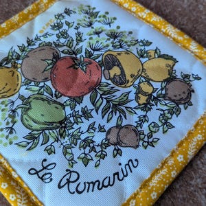 May include: A square, white pot holder with a yellow floral border. The center features a colorful illustration of various vegetables and the text "Le Romarin" in black script.