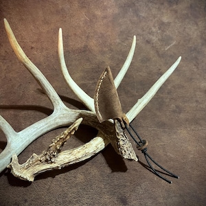 May include: A small, brown leather knife with a stitched edge and black cord is displayed against a backdrop of deer antlers. The antlers are a light, off-white color, and the background is a textured brown.