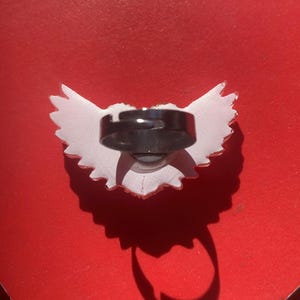 May include: A ring featuring a white, wing-shaped decorative element attached to a dark, adjustable metal band. The wing design has a jagged, feathered edge and is positioned horizontally, with the circular band centered on the back of the piece.