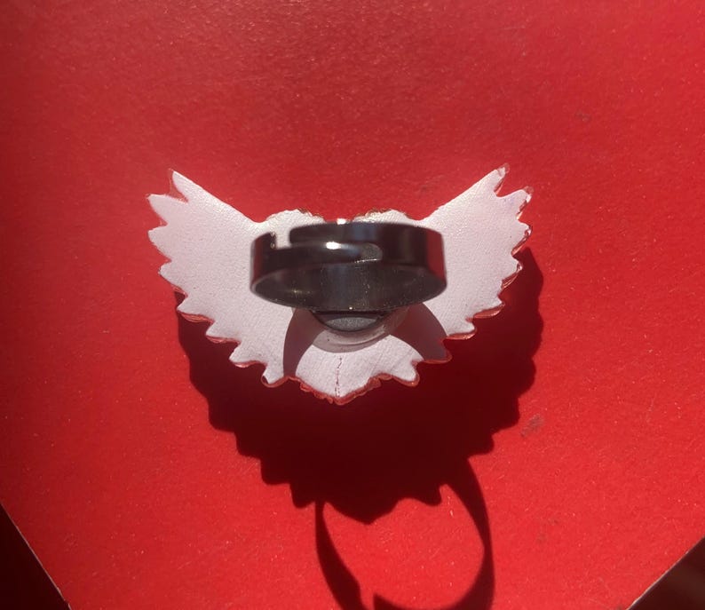 May include: A ring featuring a white, wing-shaped decorative element attached to a dark, adjustable metal band. The wing design has a jagged, feathered edge and is positioned horizontally, with the circular band centered on the back of the piece.