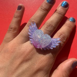 May include: A ring featuring a heart-shaped centerpiece with attached wings, crafted from a translucent material embedded with iridescent glitter. The heart is textured with small rose-like details and transitions from a clear top to a purple base.