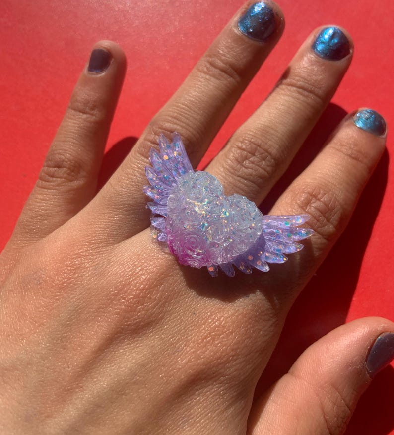 May include: A ring featuring a heart-shaped centerpiece with attached wings, crafted from a translucent material embedded with iridescent glitter. The heart is textured with small rose-like details and transitions from a clear top to a purple base.