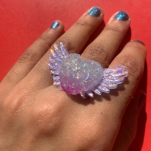 May include: A ring featuring a central heart shape with outstretched wings, crafted from a translucent, iridescent material with embedded glitter. The heart displays a gradient of light purple and pink tones, while the wings have a shimmering finish.