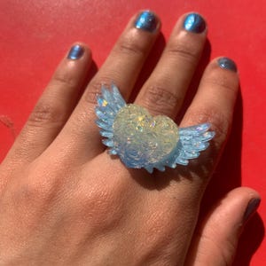 May include: A ring featuring a central heart shape adorned with a raised rose pattern, flanked by two translucent light blue wings. The heart has a pale, iridescent finish with subtle glitter, while the wings have a clear, glossy blue tint.