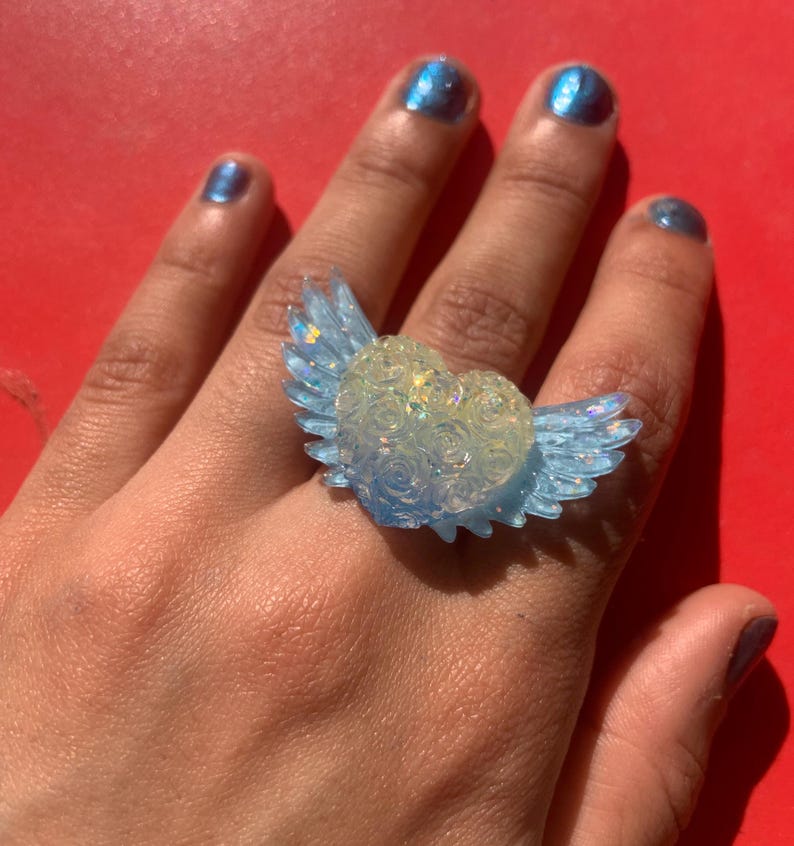 May include: A ring featuring a central heart shape adorned with a raised rose pattern, flanked by two translucent light blue wings. The heart has a pale, iridescent finish with subtle glitter, while the wings have a clear, glossy blue tint.