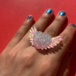 May include: A ring featuring a central heart shape adorned with a textured rose pattern, flanked by two iridescent, light pink wings. The heart and wings exhibit a translucent, glittery finish that catches the light, creating a shimmering effect.