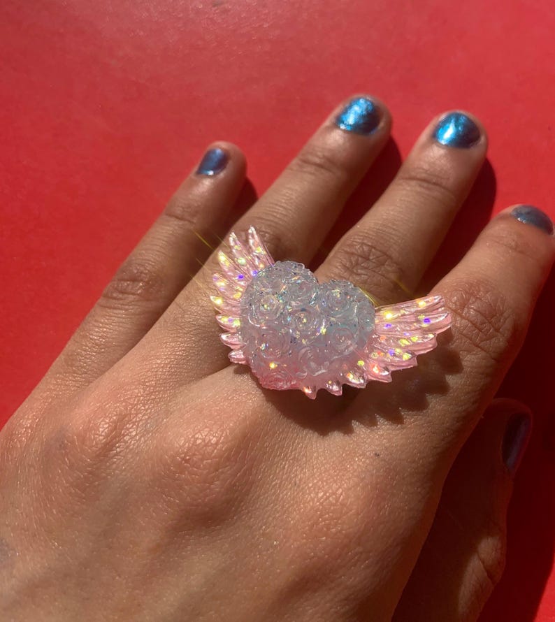 May include: A ring featuring a central heart shape adorned with a textured rose pattern, flanked by two iridescent, light pink wings. The heart and wings exhibit a translucent, glittery finish that catches the light, creating a shimmering effect.