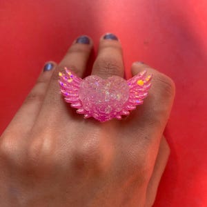 May include: A ring featuring a central heart shape with textured rose details and two outstretched wings on either side. The piece is crafted from a translucent, iridescent pink material that catches the light, creating a shimmering effect across the surface.