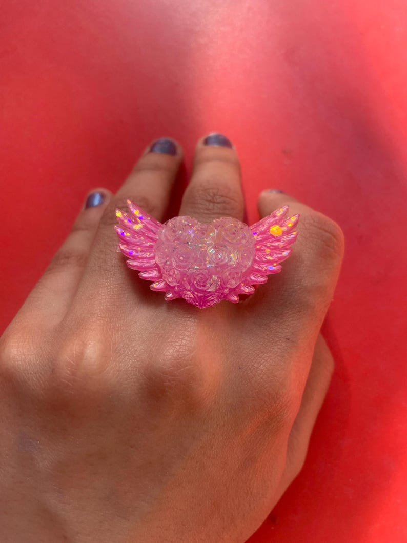 May include: A ring featuring a central heart shape with textured rose details and two outstretched wings on either side. The piece is crafted from a translucent, iridescent pink material that catches the light, creating a shimmering effect across the surface.