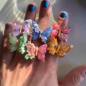 May include: A collection of six rings featuring butterfly designs in various colours including purple, blue, pink, green, orange, and yellow. Each butterfly is adorned with small floral accents on the wings, creating a detailed and colourful set of jewellery.