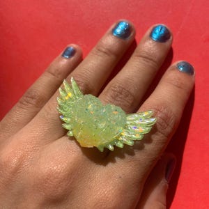 May include: A ring featuring a central heart shape with outstretched wings on both sides. The piece is crafted from a translucent, pale yellow material with an iridescent, glittery finish that catches the light, creating a shimmering effect.