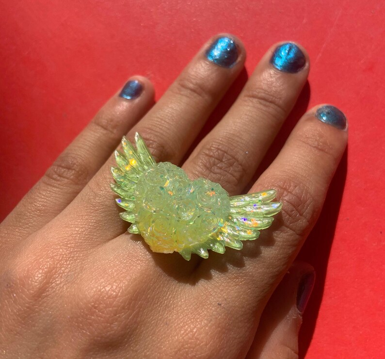 May include: A ring featuring a central heart shape with outstretched wings on both sides. The piece is crafted from a translucent, pale yellow material with an iridescent, glittery finish that catches the light, creating a shimmering effect.