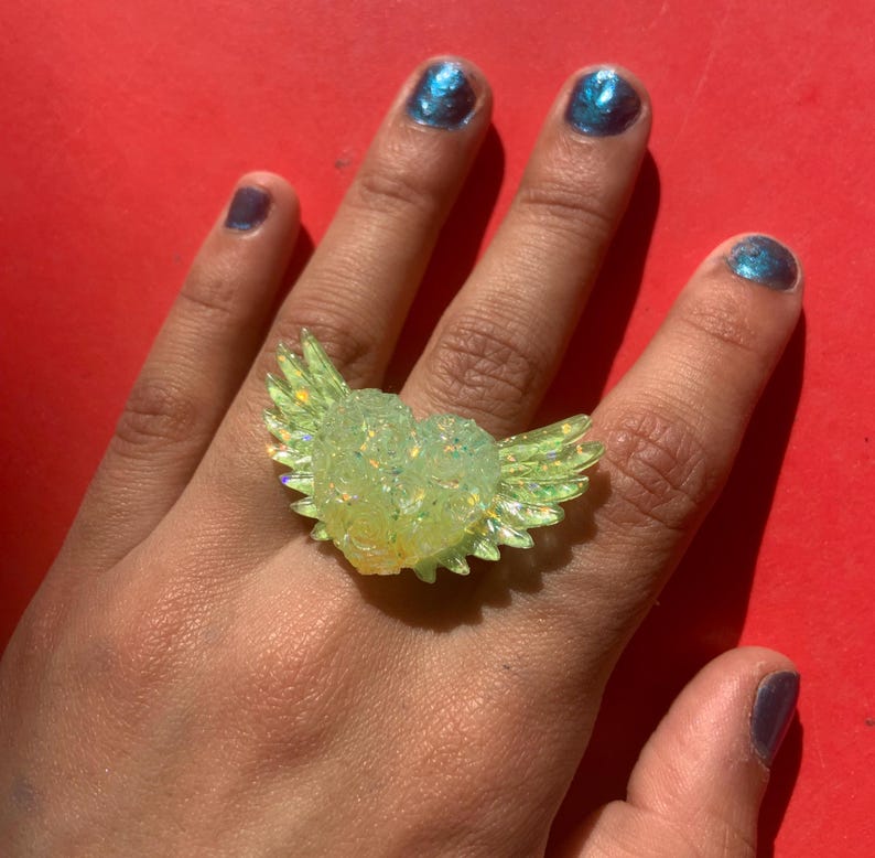 May include: A ring featuring a translucent, pale yellow heart-shaped centerpiece with iridescent glitter accents and two stylized wings extending from the sides. The design is crafted from a glossy material that catches the light, creating a shimmering effect across the surface of the heart and the feathered wing details.