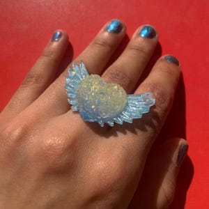 May include: A ring featuring a central heart-shaped element with a textured rose pattern, flanked by two translucent light blue wings. The heart has a pale, iridescent finish that catches the light, while the wings have a clear, glossy appearance.