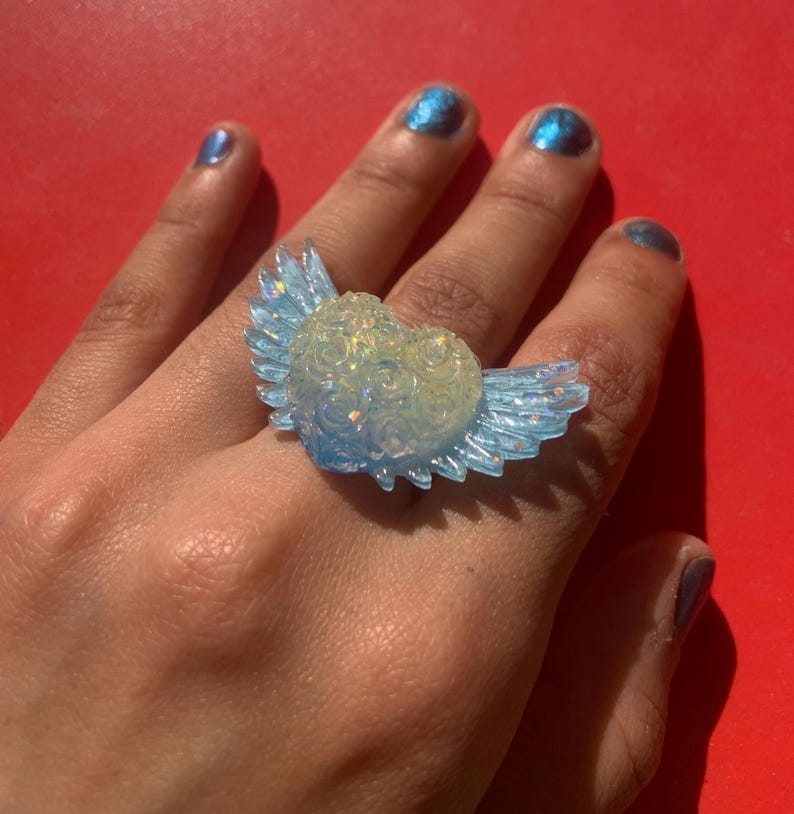 May include: A ring featuring a central heart-shaped element with a textured rose pattern, flanked by two translucent light blue wings. The heart has a pale, iridescent finish that catches the light, while the wings have a clear, glossy appearance.