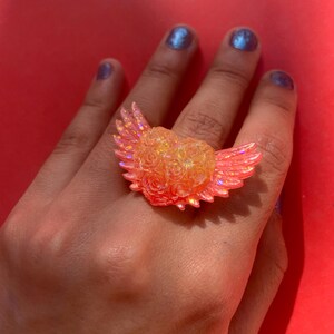 May include: A ring featuring a central heart shape composed of small, textured rose motifs, flanked by two iridescent, translucent orange wings. The entire piece has a shimmering, glittery finish that catches the light, creating a bright, warm glow.