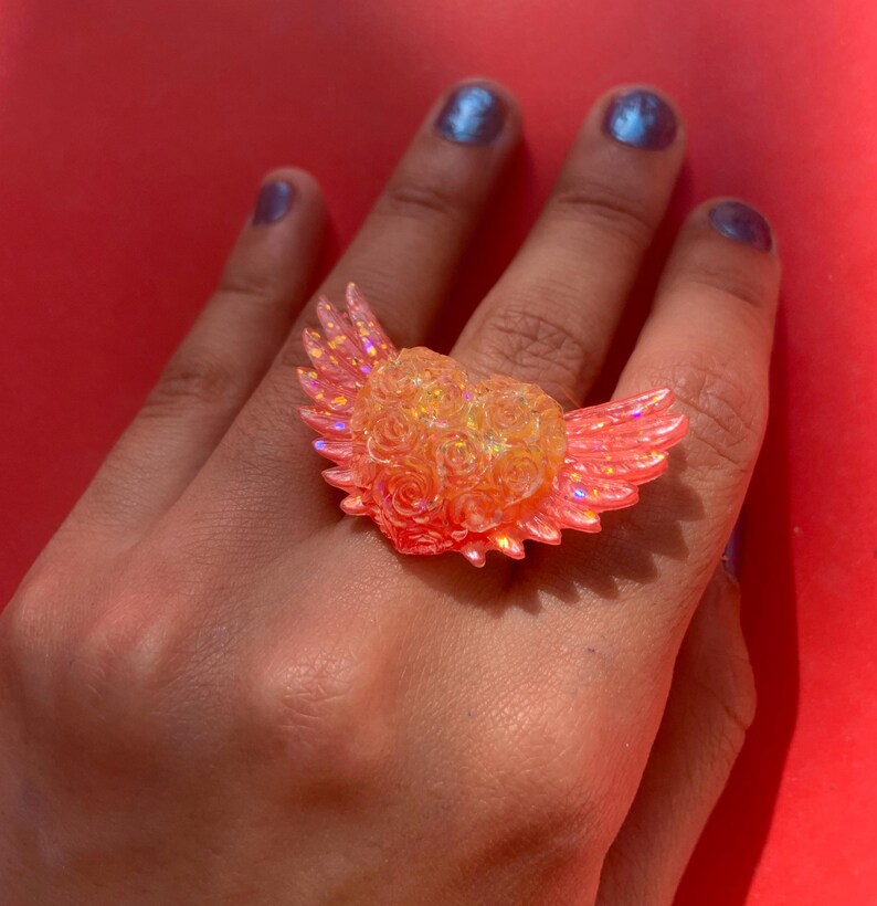 May include: A ring featuring a central heart shape composed of small, textured rose motifs, flanked by two iridescent, translucent orange wings. The entire piece has a shimmering, glittery finish that catches the light, creating a bright, warm glow.