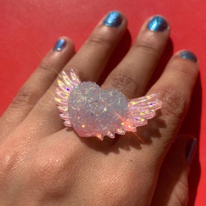 May include: A ring featuring a heart-shaped centerpiece adorned with small sculpted roses and iridescent wings extending from the sides. The material is translucent and light-catching, displaying a shimmering effect with pink and blue highlights.