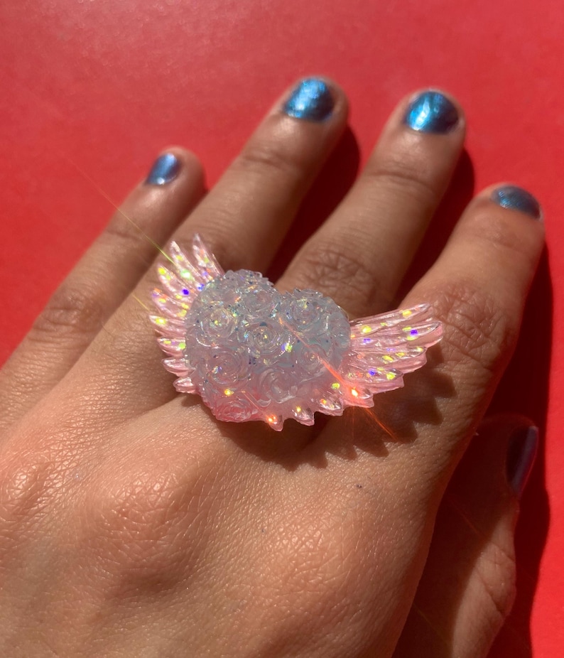 May include: A ring featuring a heart-shaped centerpiece adorned with small sculpted roses and iridescent wings extending from the sides. The material is translucent and light-catching, displaying a shimmering effect with pink and blue highlights.