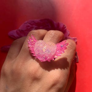 May include: A ring featuring a central heart shape adorned with a textured rose pattern and iridescent finish, flanked by two translucent pink wings. The heart and wings are crafted from a glossy material that reflects light with a shimmering effect.