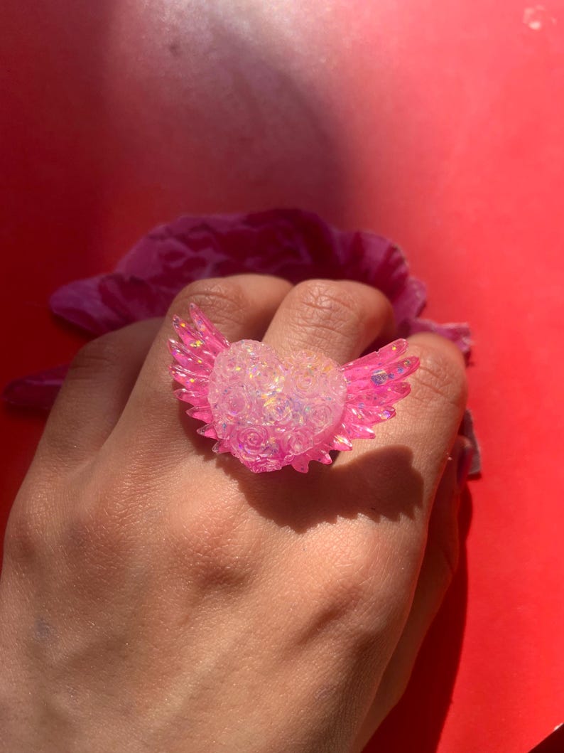 May include: A ring featuring a central heart shape adorned with a textured rose pattern and iridescent finish, flanked by two translucent pink wings. The heart and wings are crafted from a glossy material that reflects light with a shimmering effect.
