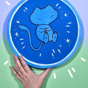 May include: A circular blue painting depicting a light blue cartoon character with a long tail, seated with hands clasped. White starbursts and a white border surround the character. The painting is held by a hand against a green and blue background.