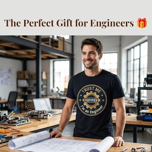 Puede incluir: Una camiseta negra de manga corta con un gráfico circular dorado y negro que incluye un icono de engranaje y el texto "TRUST ME I'M AN ENGINEER" y "CERTIFIED" impreso en la parte delantera. La camiseta se lleva en un espacio de trabajo.